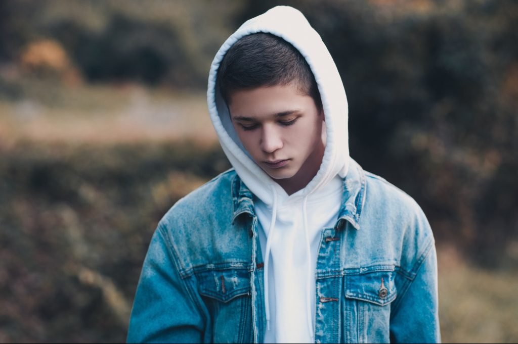 Sad teen boy 14-16 year old wearing white hoodie and denim jacket in dark outdoors. Depressed mood.